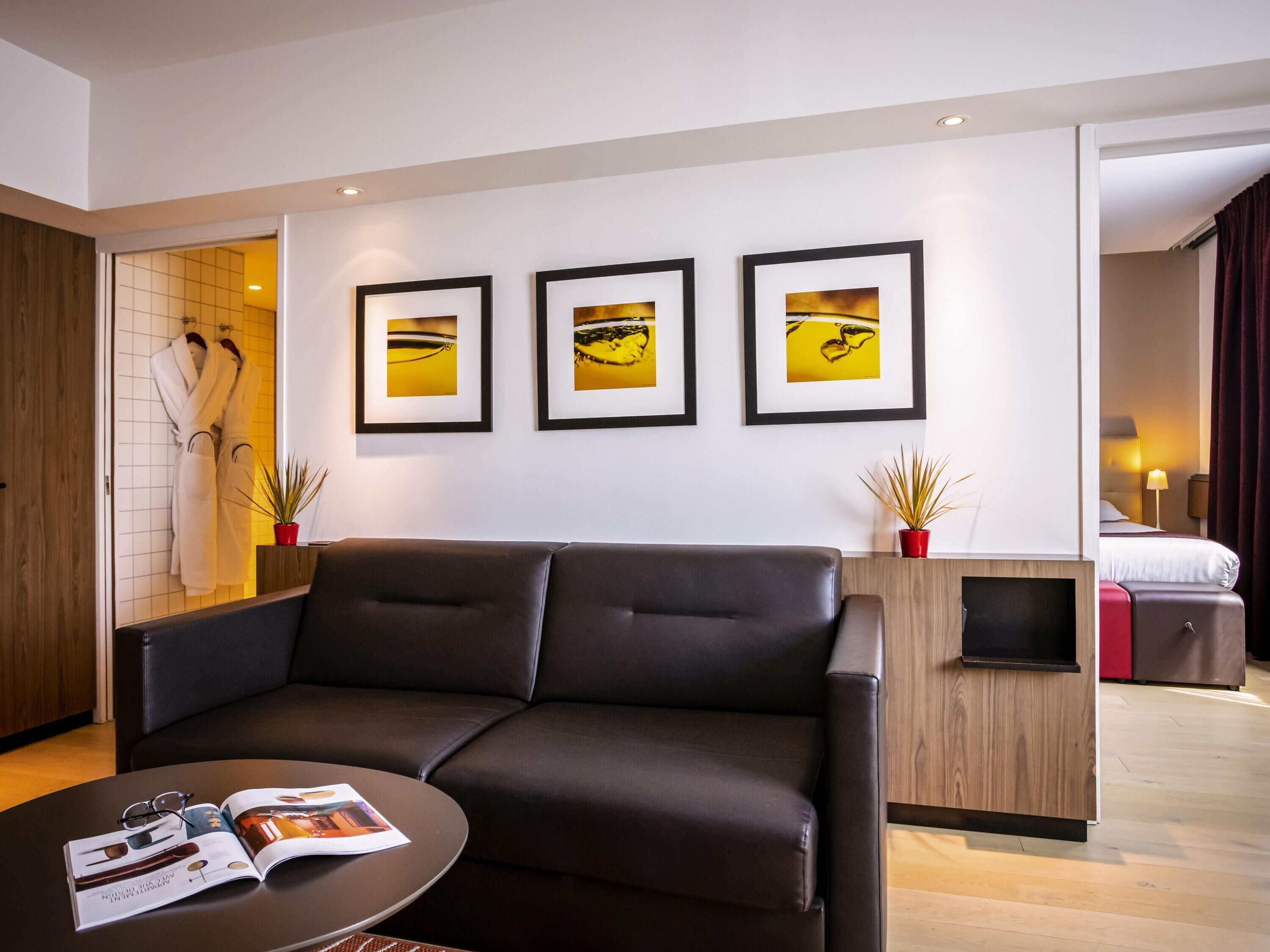 Une chambre d’hôtel moderne avec un canapé en cuir, une petite table basse sur laquelle repose un magazine, un meuble en bois et trois tableaux encadrés accrochés au mur.