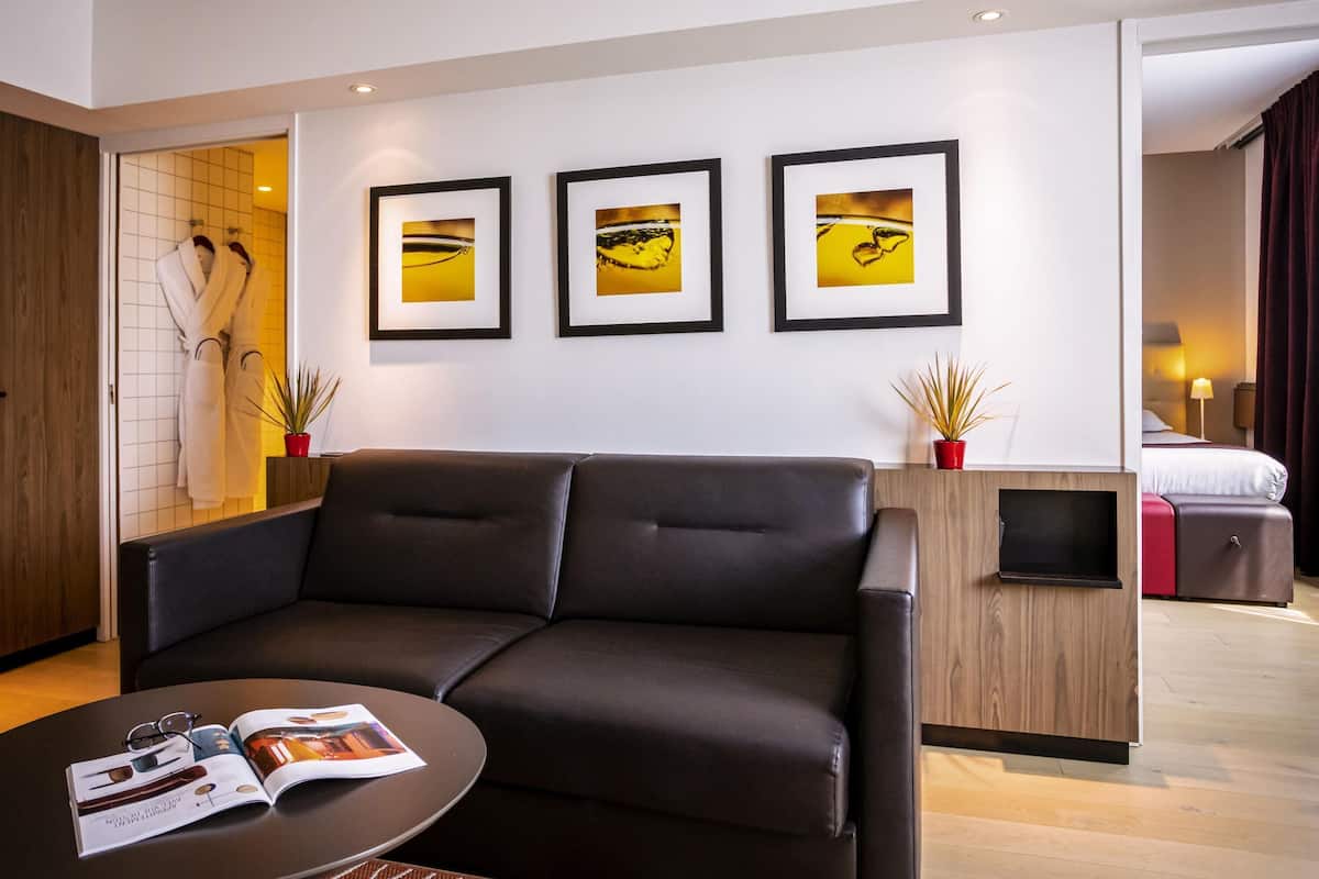 A modern hotel room with a leather sofa, a small coffee table with a magazine, a wooden cabinet, and three framed pictures on the wall.