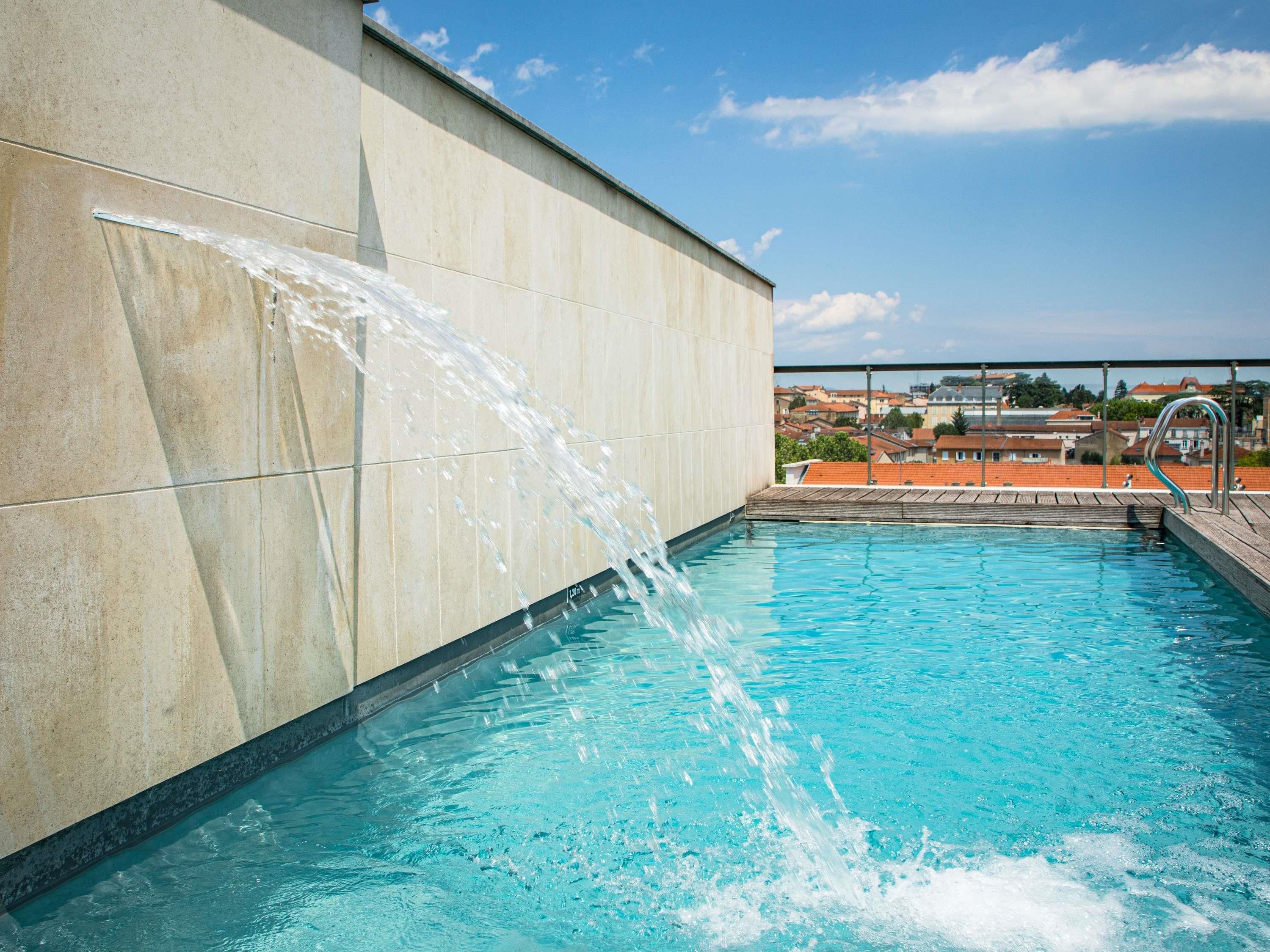 Una piscina con agua cayendo por una pared, rodeada de un suelo de baldosas y un cielo despejado.