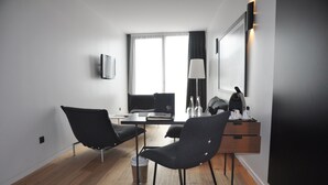 A modern hotel room with a wooden floor, a desk with a chair, a flat-screen TV mounted on the wall, and a window with curtains.