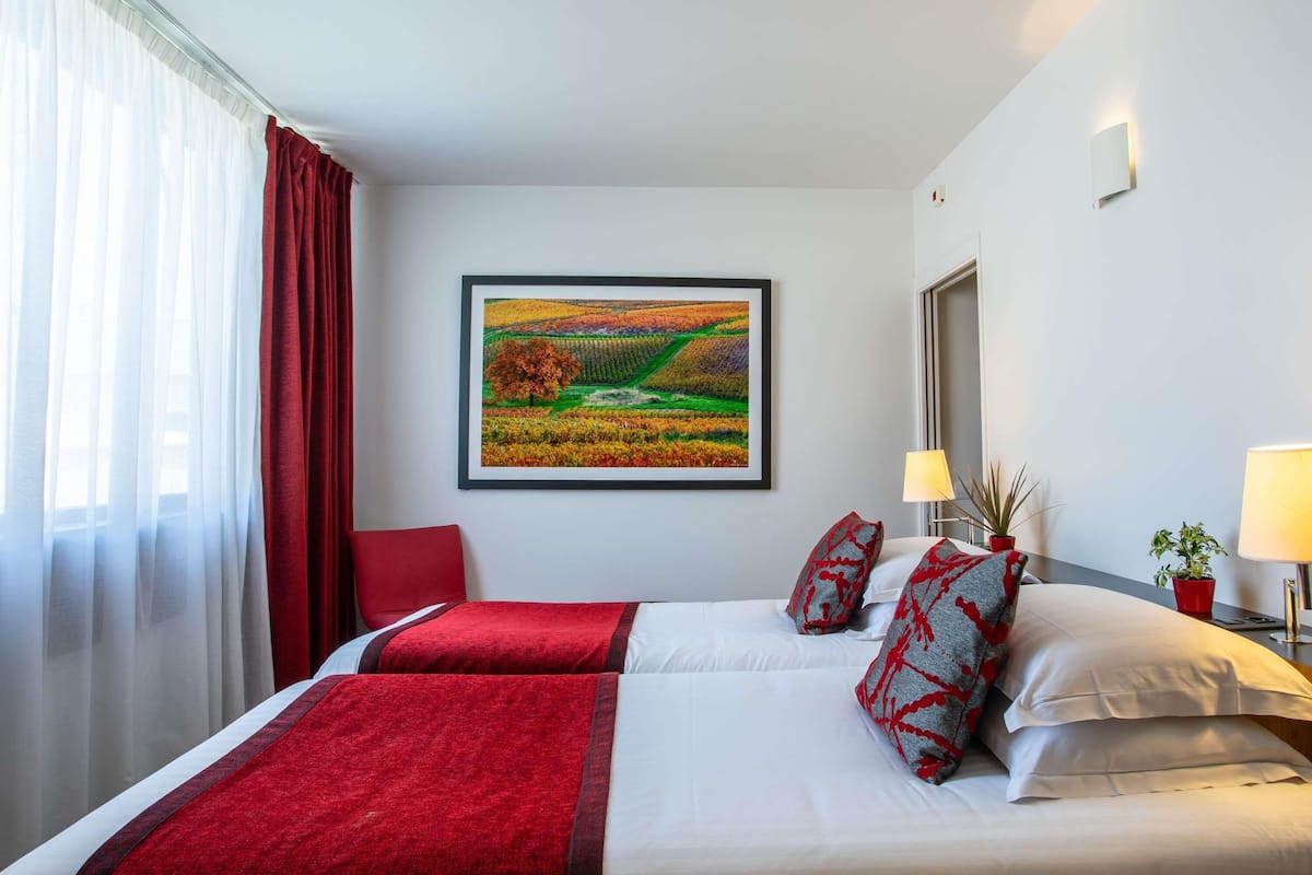 A hotel room with a bed, red curtains, a framed landscape painting, a bedside table with a lamp, and a potted plant.