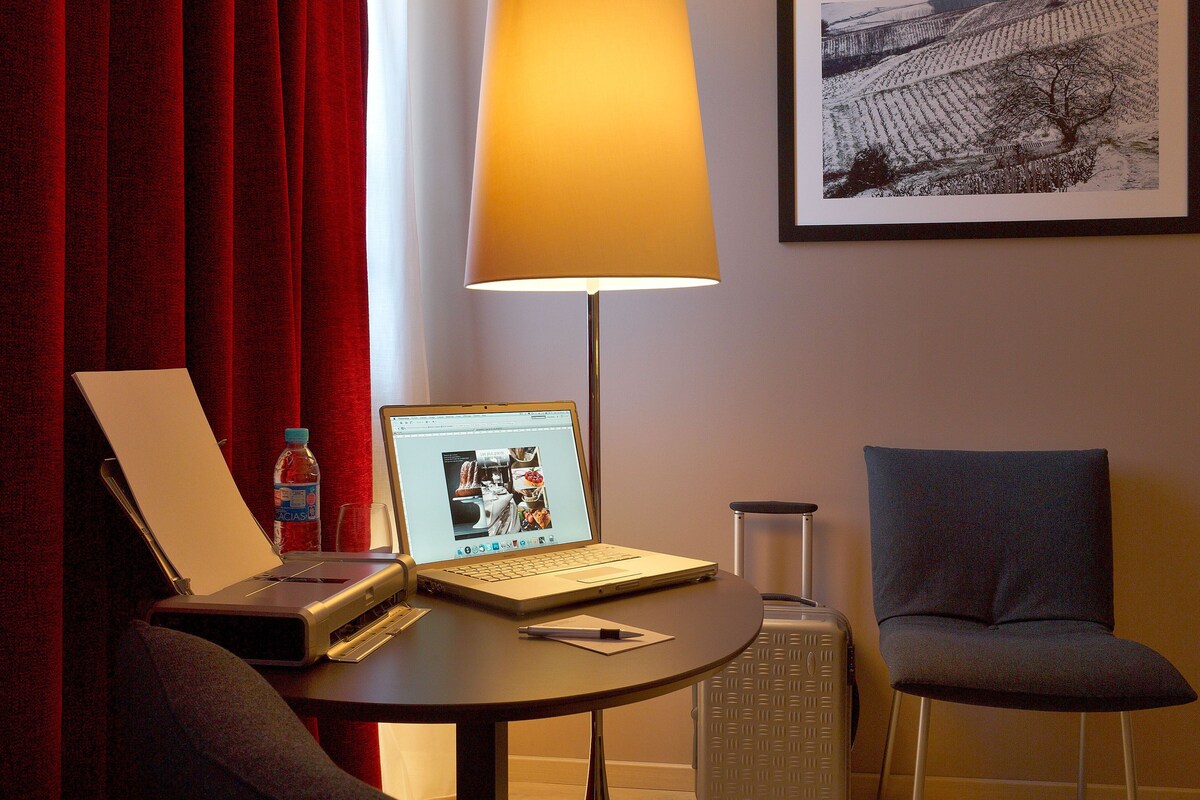 A hotel room with a laptop on a table, a lamp, a chair, and a framed picture on the wall.