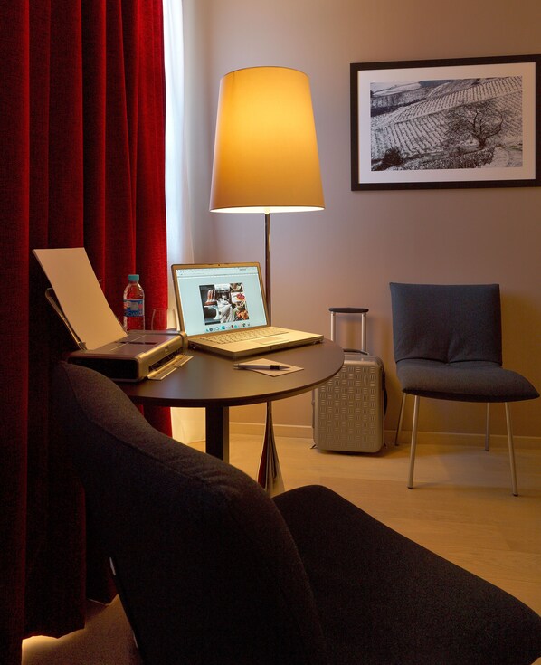 A hotel room with a laptop on a table, a lamp, a chair, and a framed picture on the wall.