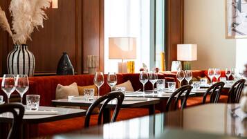 A restaurant with a long dining table, multiple chairs, and a sofa. There are several wine glasses and a vase with dried plants on the table.