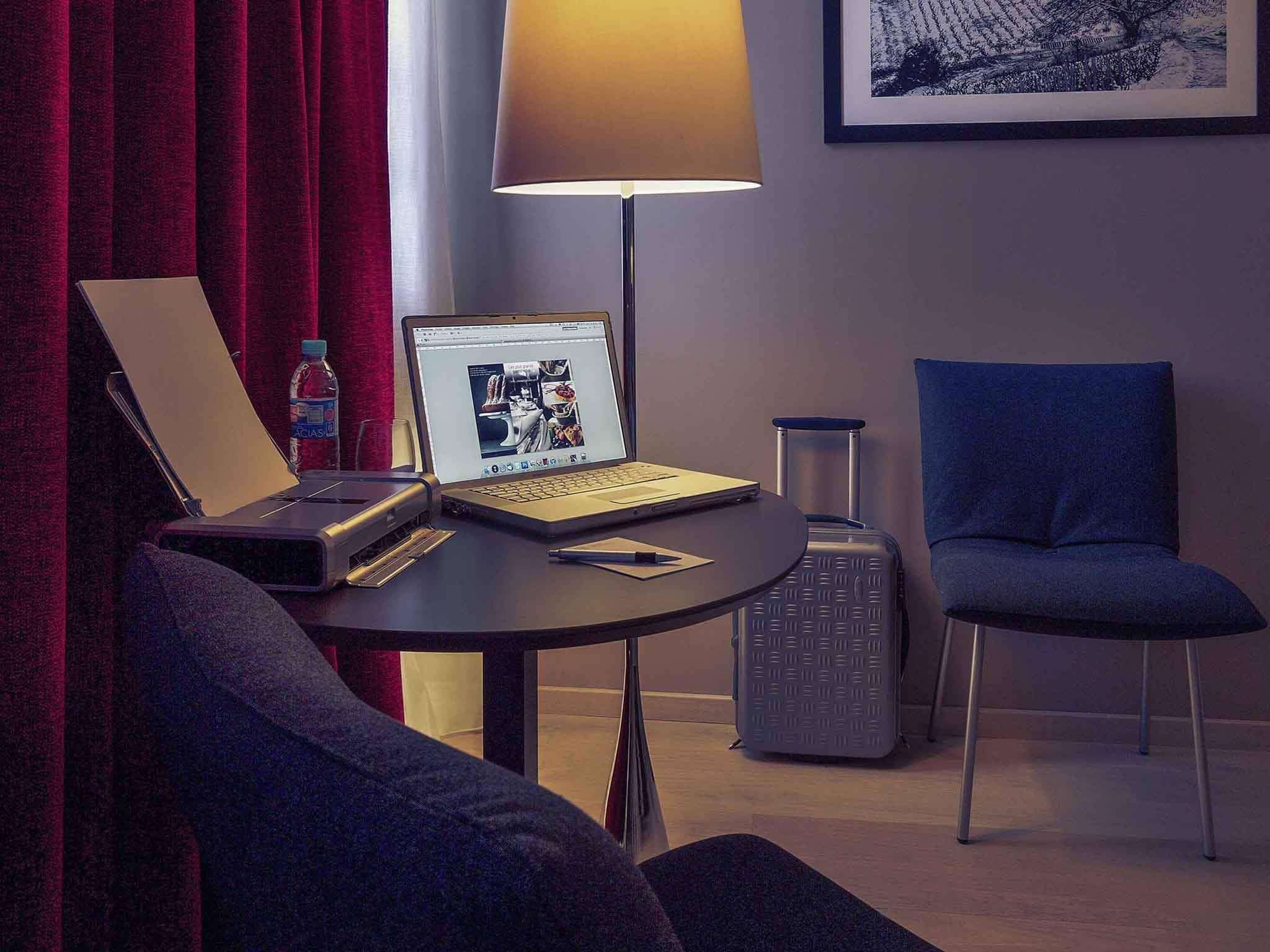 Una habitación de hotel con una computadora portátil sobre una mesa, una lámpara, una silla y una maleta.