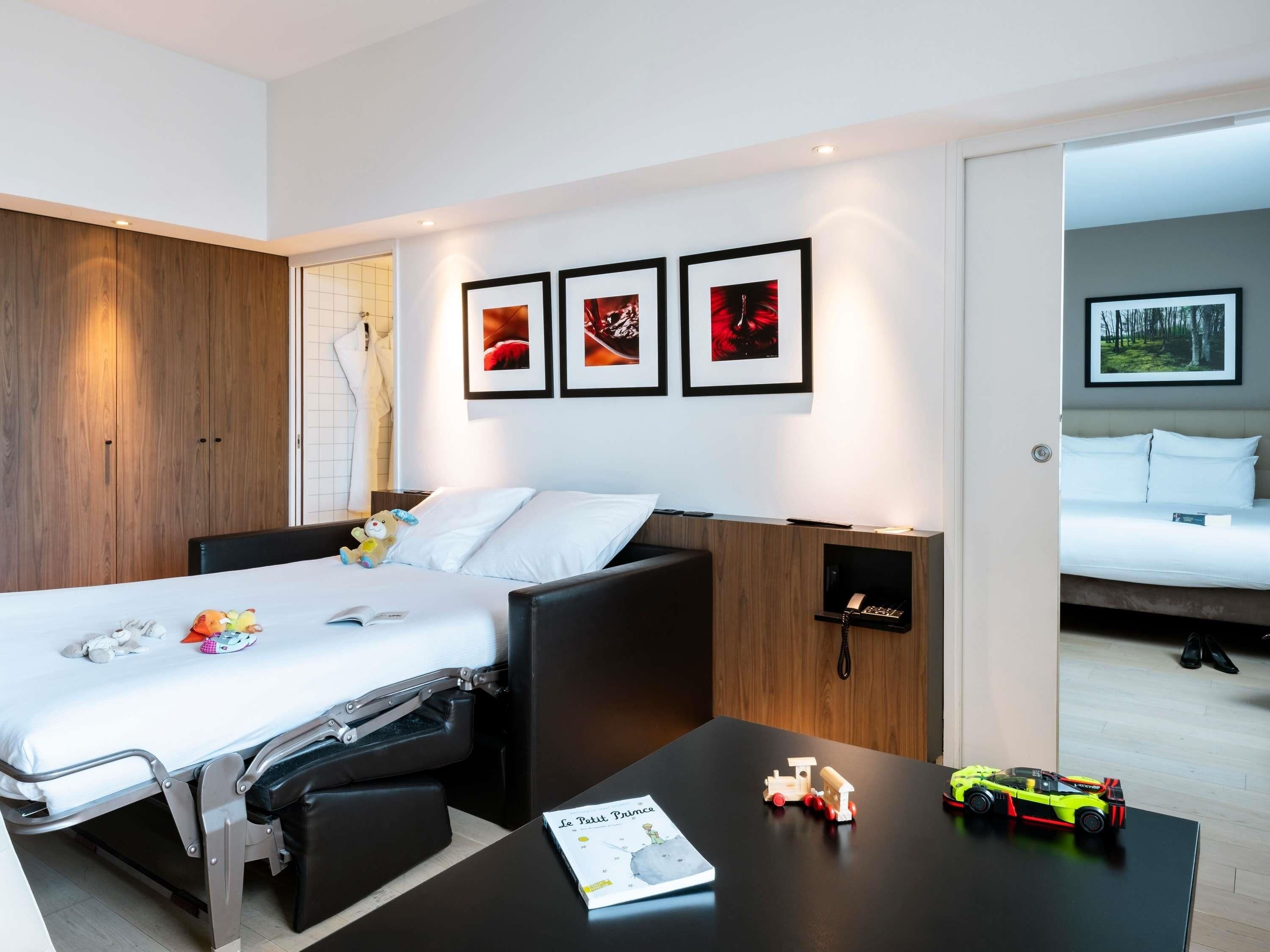 A modern hotel room with a convertible bed, a small desk, and a bookshelf.