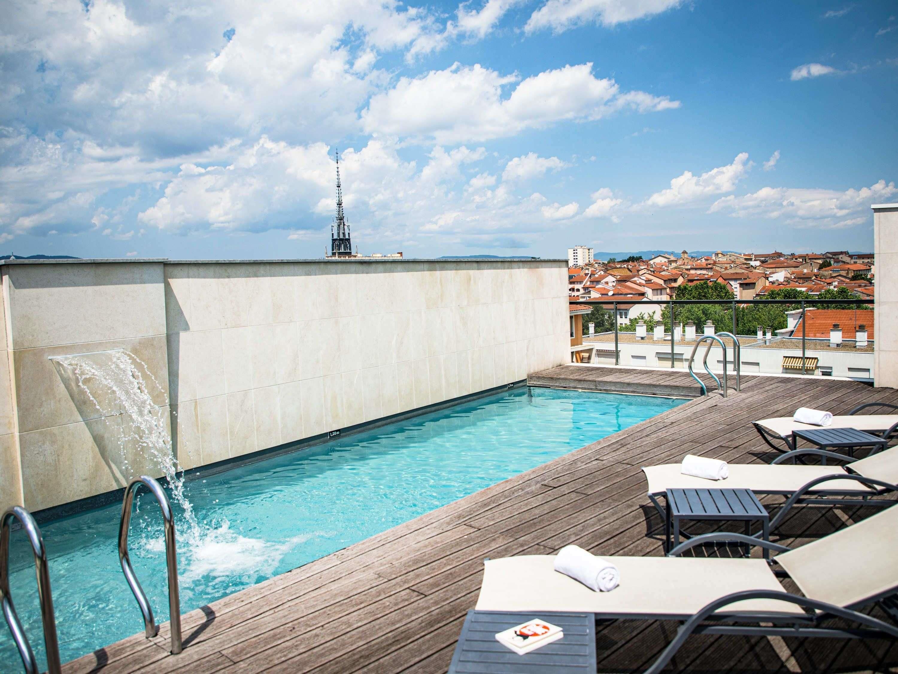 Une piscine sur le toit, avec une terrasse en bois, des chaises longues et une vue sur le paysage urbain.