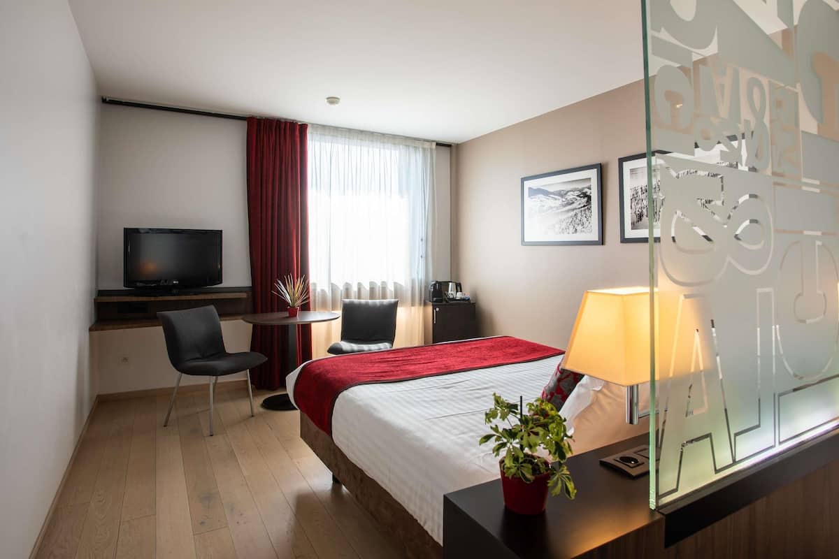 A hotel room with a bed, a desk with a television, a chair, a small table, a lamp, and a potted plant.