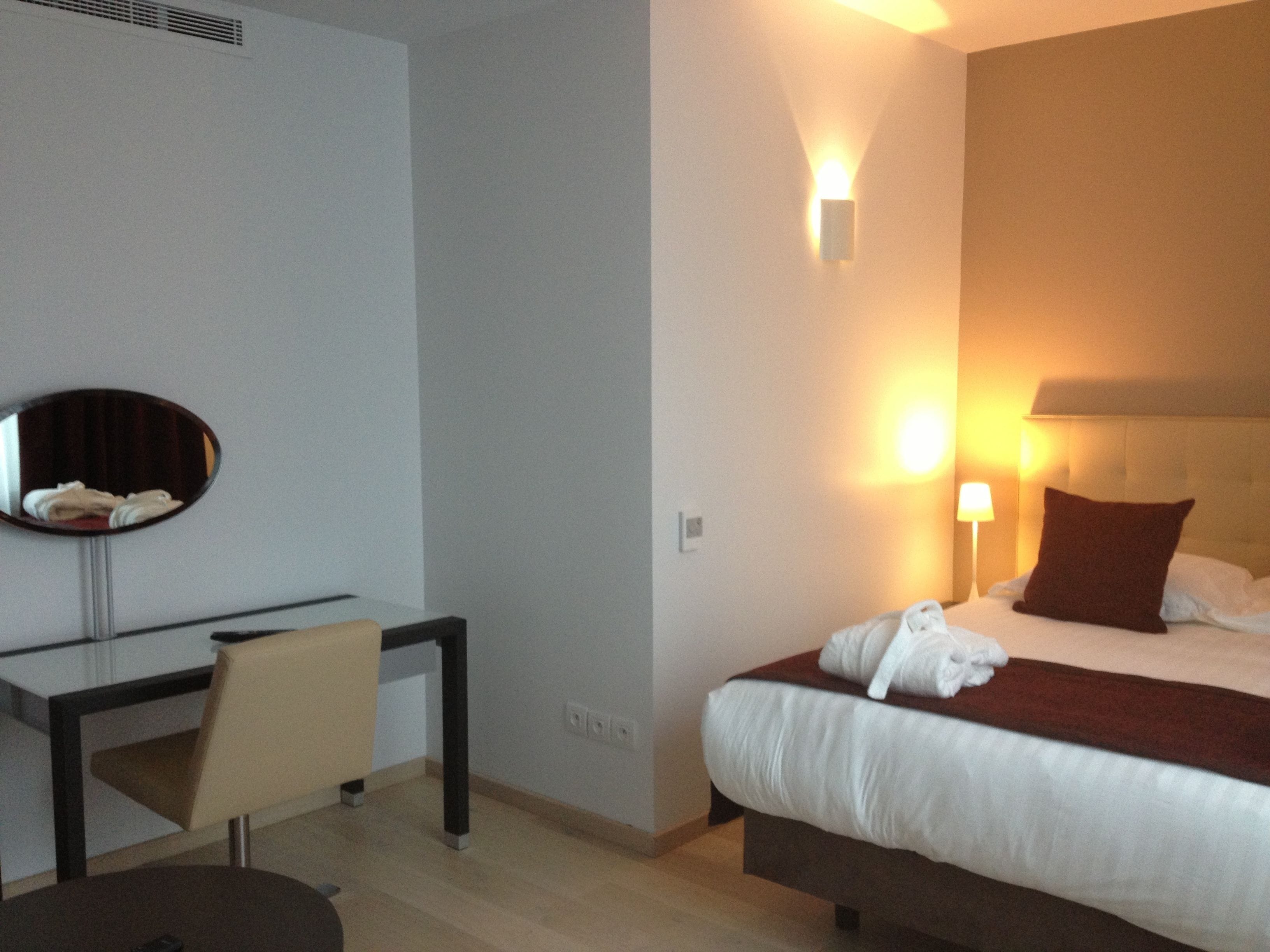 A hotel room with a bed, a desk, a chair, a mirror, and a wall-mounted light fixture.