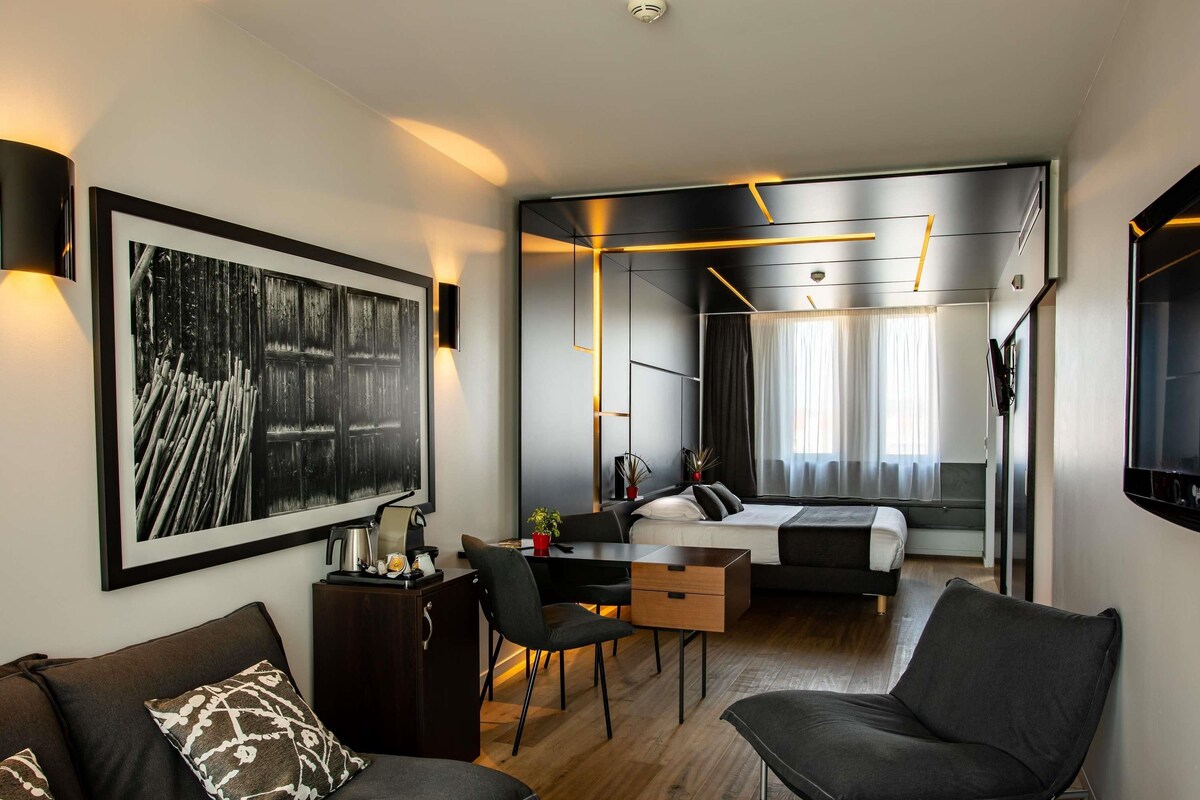 A modern hotel room with a large bed, a desk, a sofa, and a dining area.