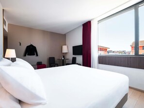 A hotel room with a bed, a desk, a chair, a TV, and a window with a view of buildings. - Hotel Mercure Villefranche-sur-Saone (Villefranche-sur-Saône)