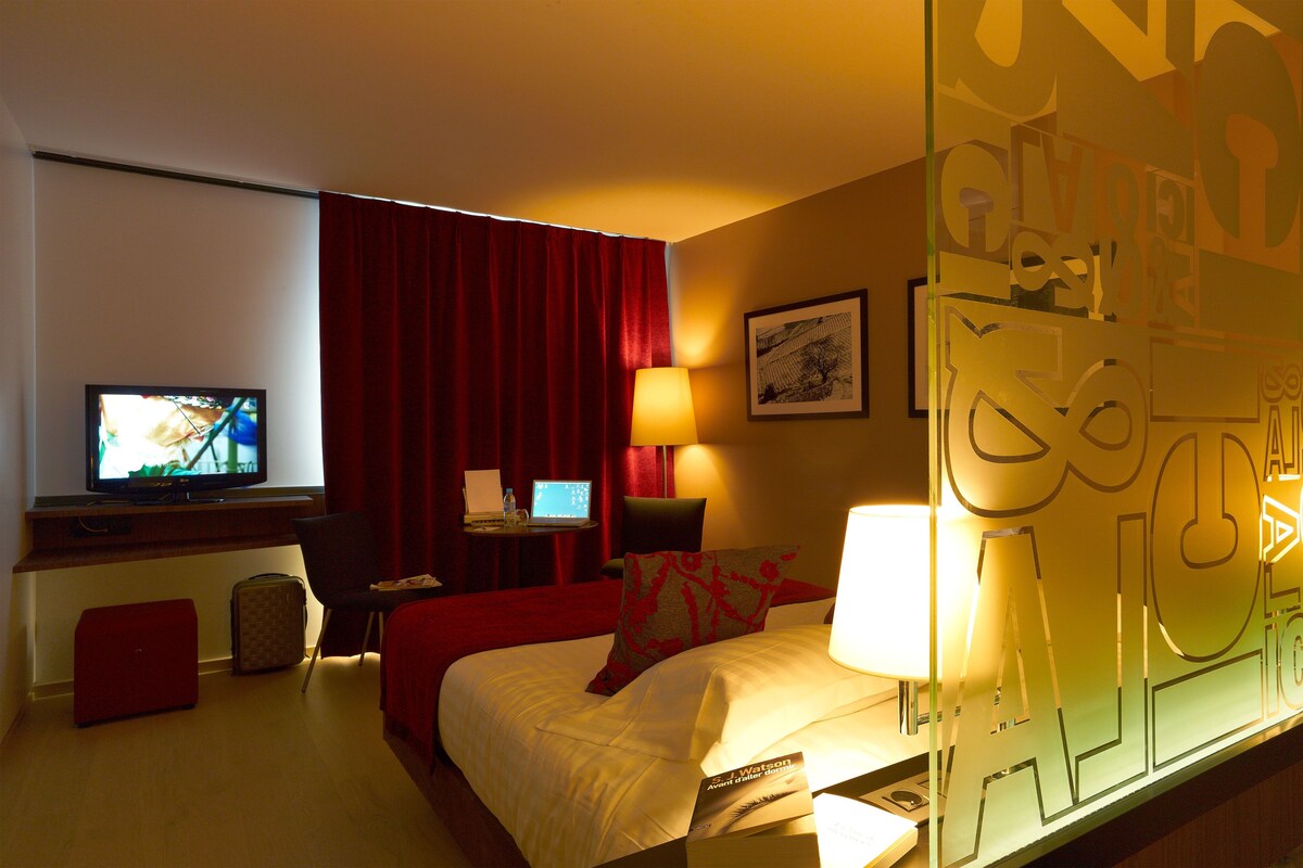 A hotel room with a bed, a desk with a computer, a television, and a red curtain.
