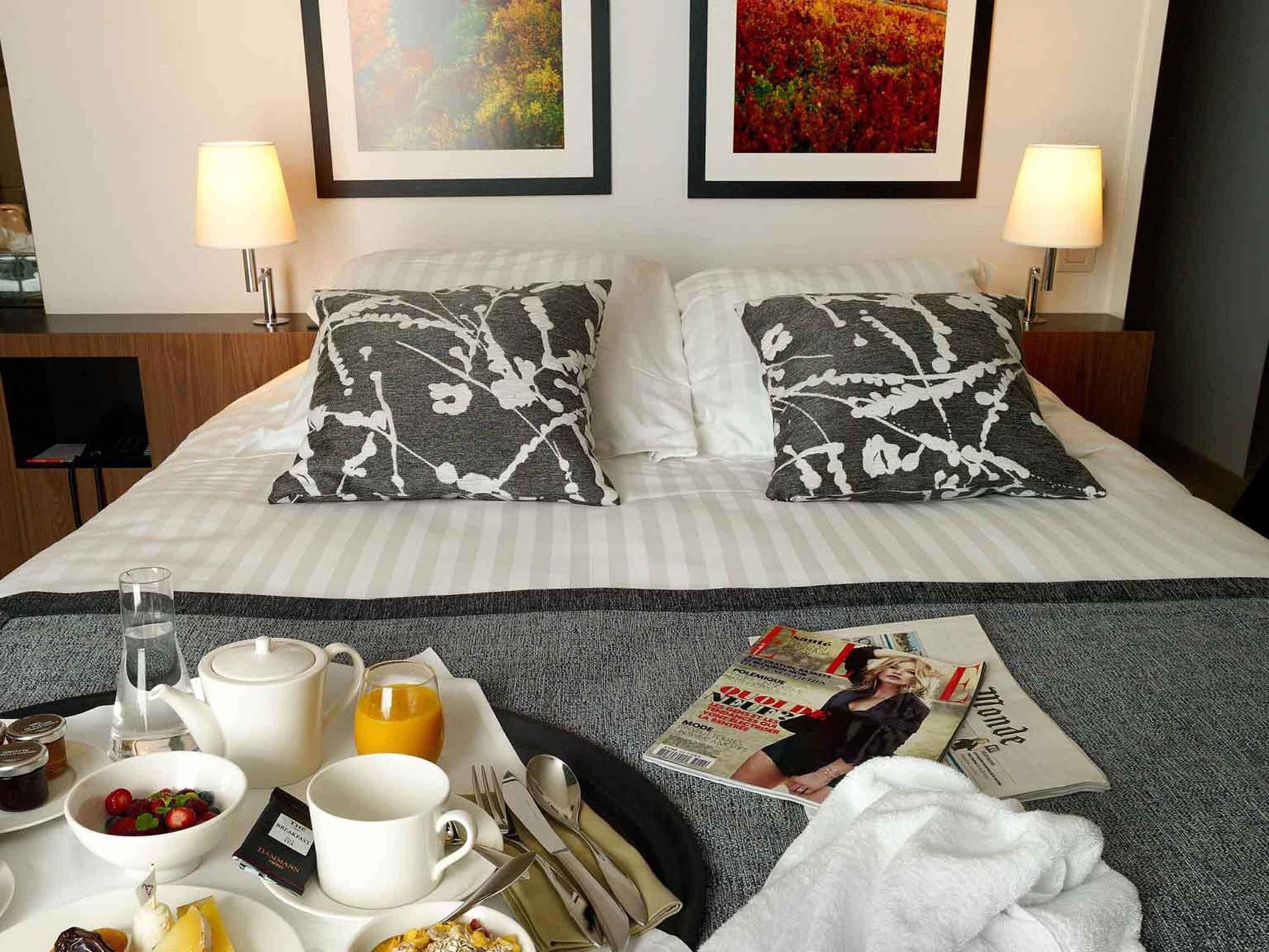 A hotel room with a neatly made bed, two pillows with a black and white abstract pattern, a tray with breakfast items, a magazine, and two framed pictures on the wall.