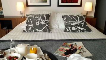 A hotel room with a neatly made bed, two pillows with a black and white abstract pattern, a tray with breakfast items, a magazine, and two framed pictures on the wall.