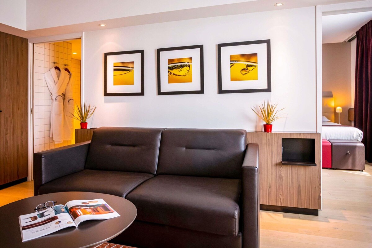 Una habitación de hotel moderna con un sofá de cuero, una mesita de café pequeña y una cama con una maleta roja. Hay tres cuadros en la pared y un armario de madera.