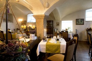 Un restaurant où les tables sont dressées pour le service, avec un décor rustique comprenant des piliers en pierre et un lustre.