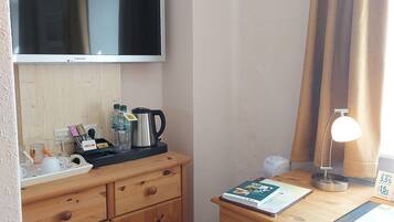 A hotel room with a wooden desk, a flat-screen TV, a coffee maker, a lamp, and a small table with a book and a menu.