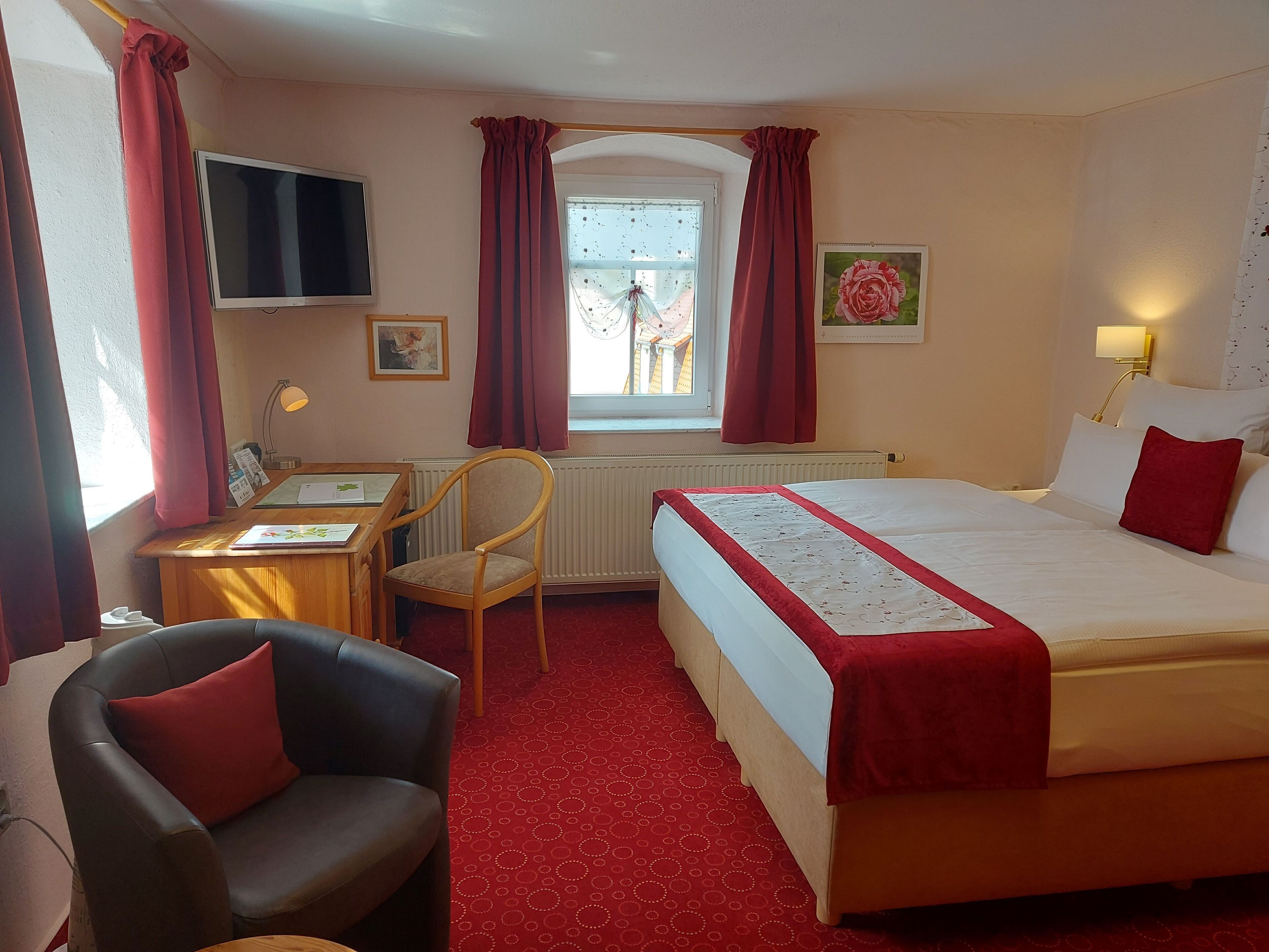 A hotel room with a bed, a desk, a chair, a TV, and a window with curtains.