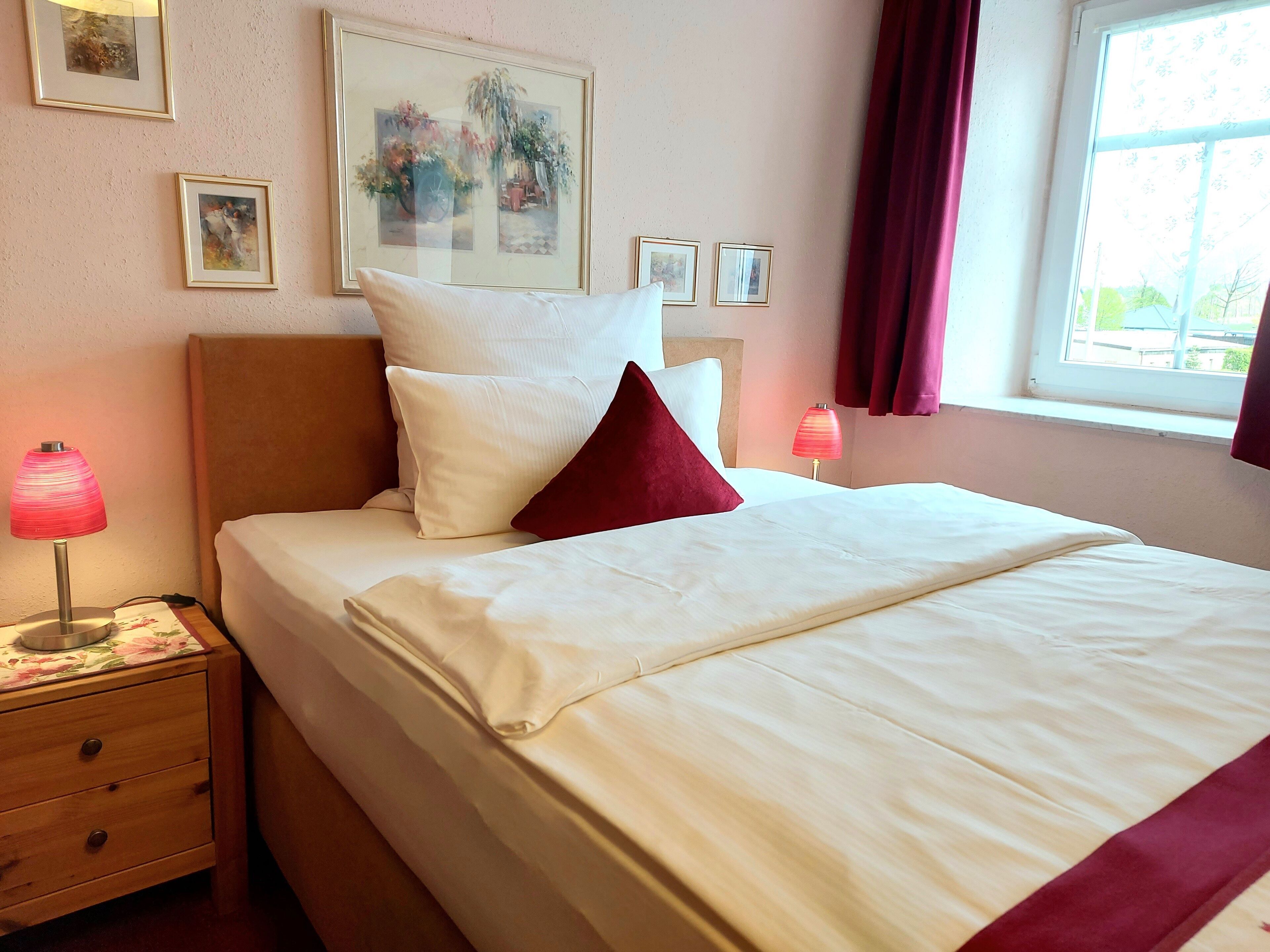 A neatly made bed with white linens and a red accent pillow, a wooden nightstand with a lamp, and a window with curtains.