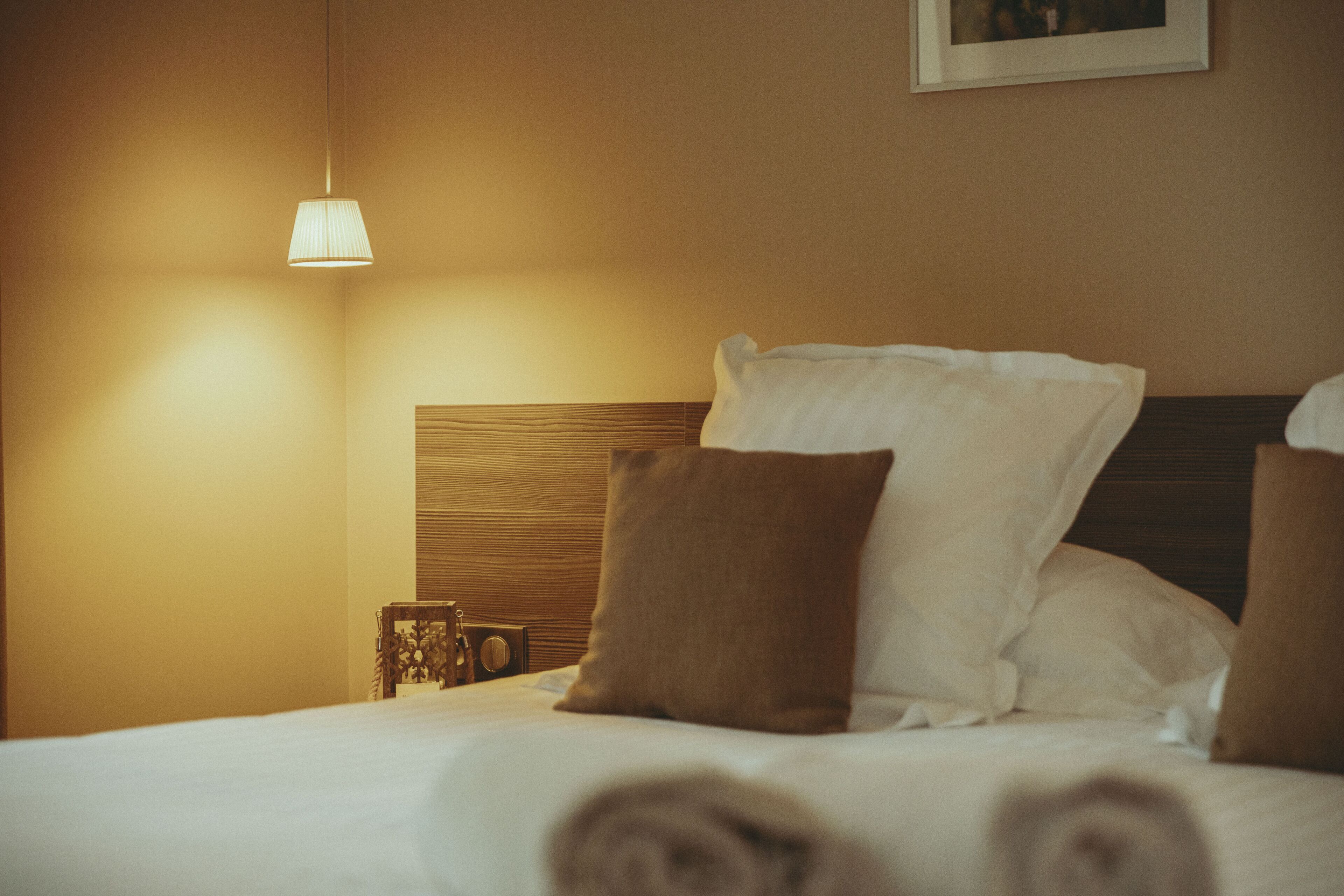 Un lit bien fait, avec des oreillers blancs et bruns, une tête de lit en bois et une table de chevet surmontée d’une lampe.