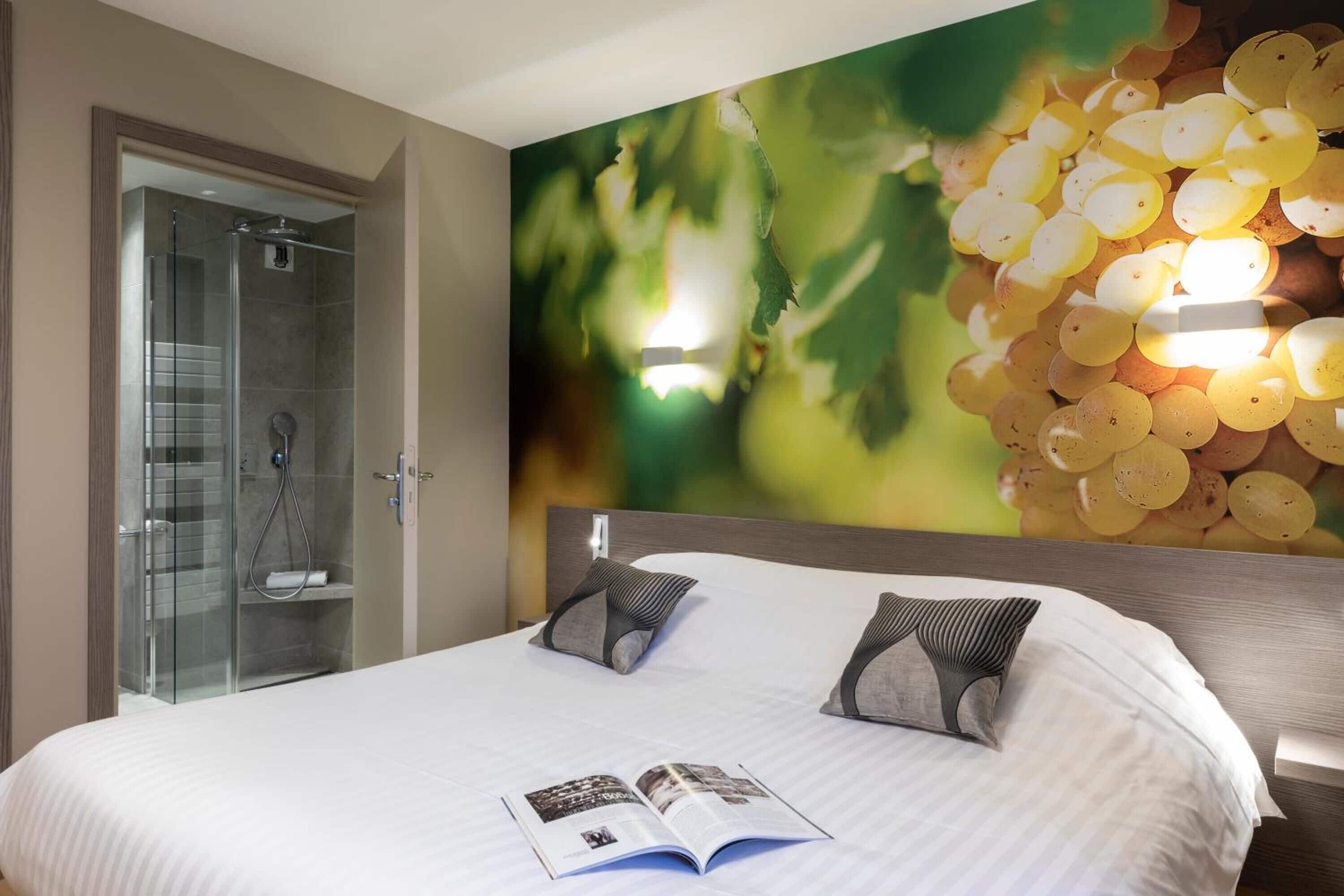 A hotel room with a large bed, a glass shower enclosure, and a wall mural of grapes.