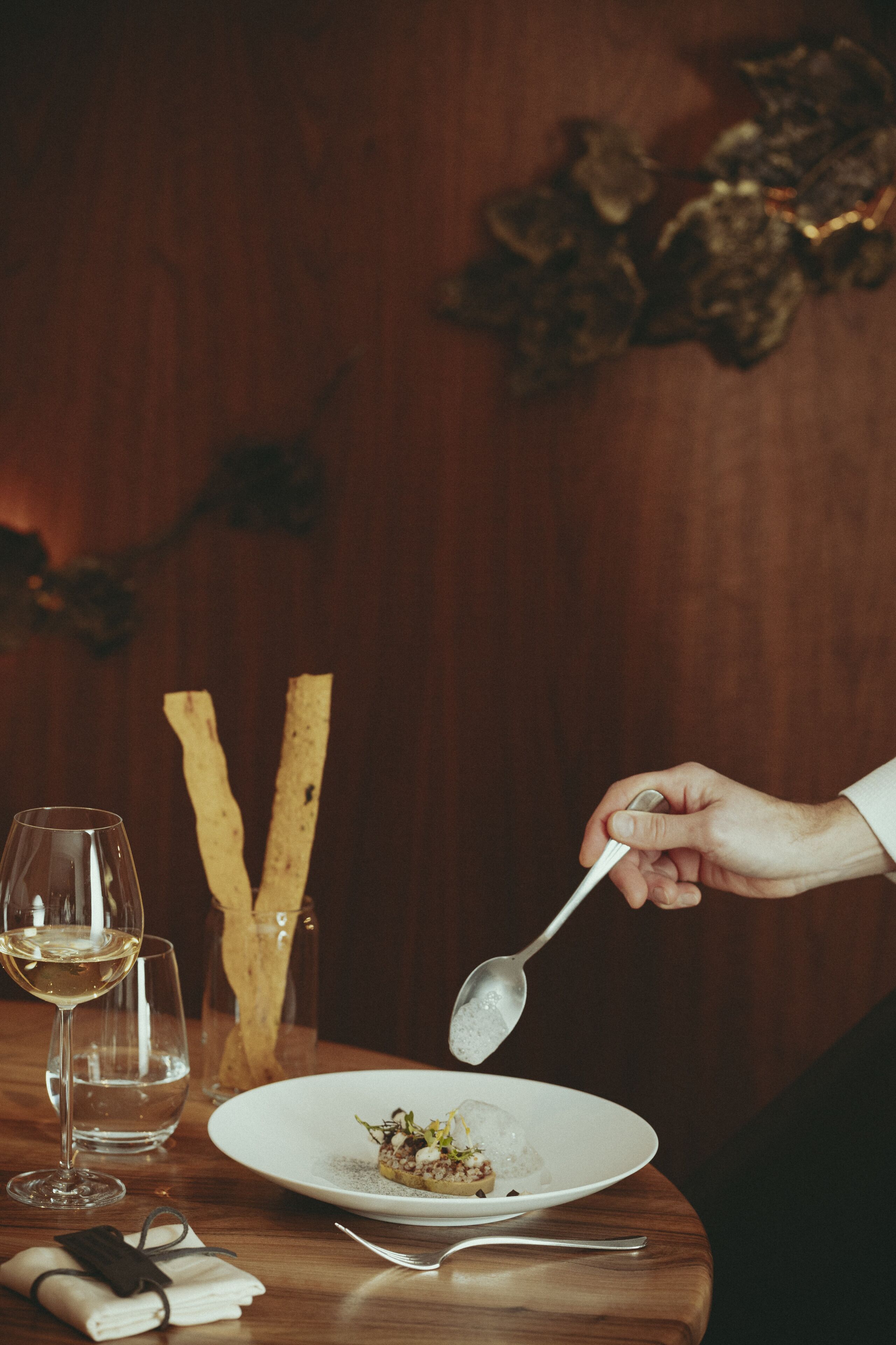 A dining table with a plate of food, a glass of white wine, and a spoon.