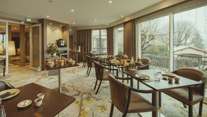 A hotel dining area with multiple tables and chairs, a large window with a view of the outside, and a plant on the side.