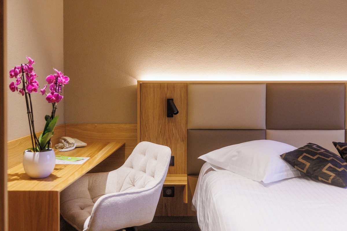 Ein Hotelzimmer mit einem Holztisch, einem weißen Sessel, einem Nachttisch, einem Bett mit weißen und gemusterten Kissen sowie einer Wandlampe.