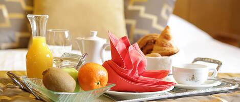 Un plateau de petit-déjeuner avec du jus d’orange, des kiwis, une orange, un croissant et une tasse de café, posé sur un lit recouvert d’une courtepointe à motifs.