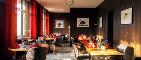 Un restaurant avec des tables et des chaises en bois, des rideaux rouges et des œuvres d’art encadrées accrochées aux murs.