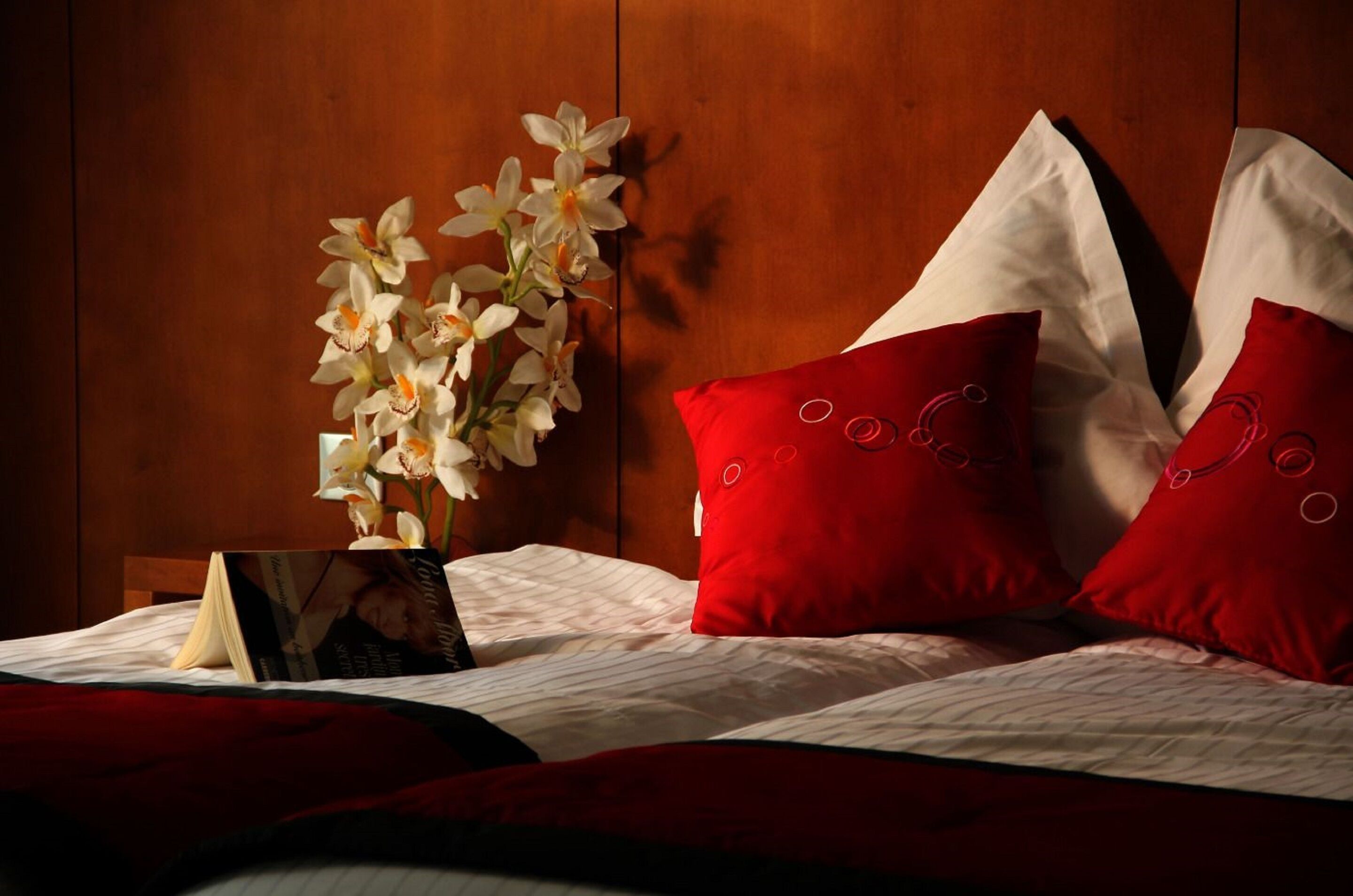 A neatly made bed with red and white pillows, a vase with white flowers, and a photo frame on the side.