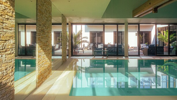 An indoor swimming pool with clear glass walls, stone pillars, and a view of outdoor seating areas.