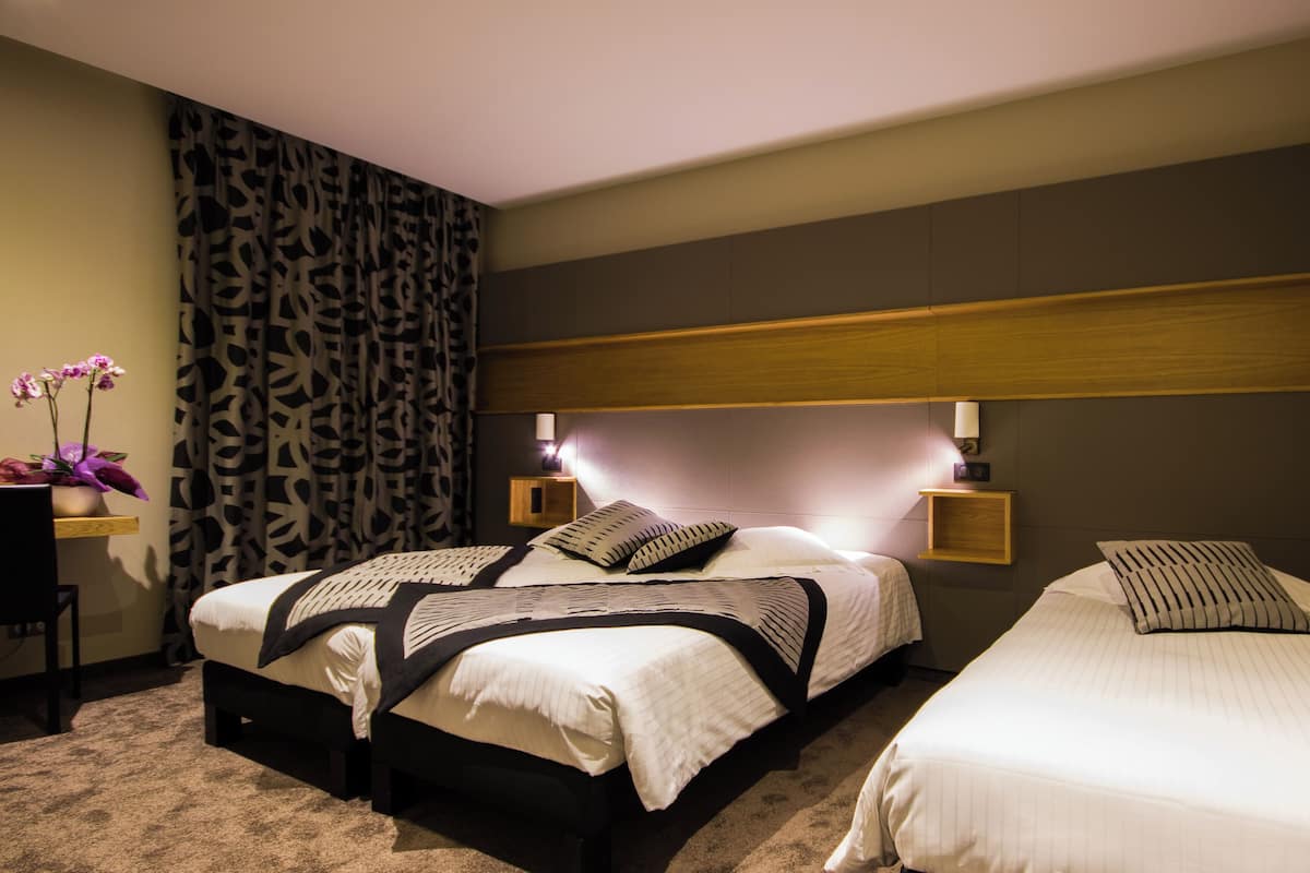 Ein Hotelzimmer mit zwei Betten, einem Nachttisch mit Lampe, einem kleinen Tisch mit Blumenvase und einem gemusterten Vorhang.
