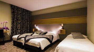 A hotel room with two beds, a nightstand with a lamp, a small table with a vase of flowers, and a patterned curtain.