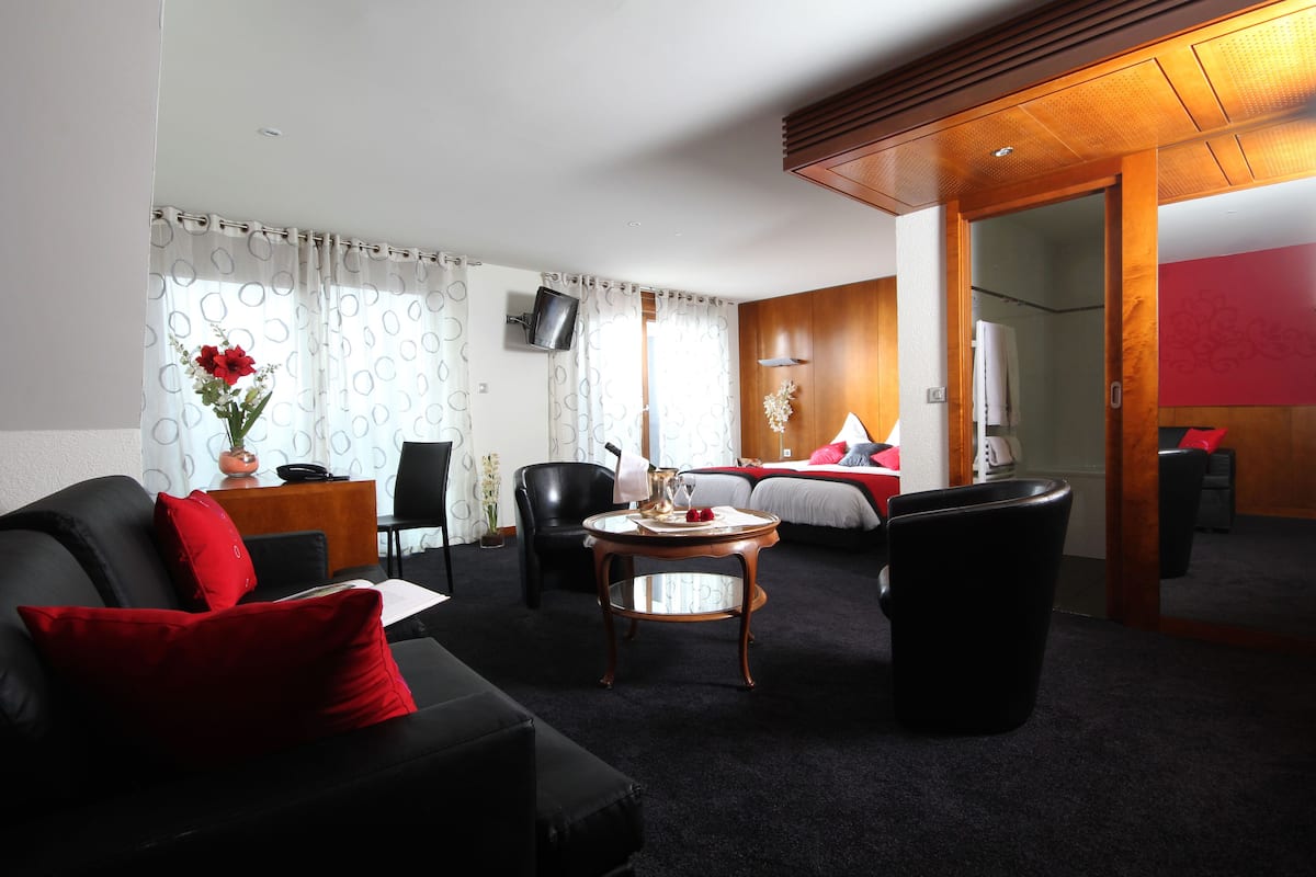 Ein Hotelzimmer mit schwarzem Ledersofa, einem Holztisch, einem Bett mit roter Bettwäsche, einem an der Wand befestigten Fernseher und einer Holzzielung.