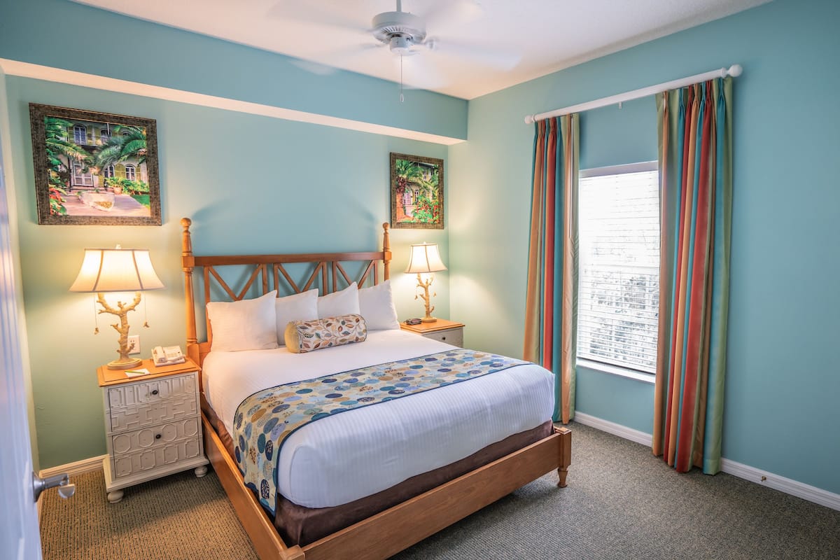 A bedroom with a wooden bed, two bedside tables with lamps, a window with curtains, and two wall art pieces.
