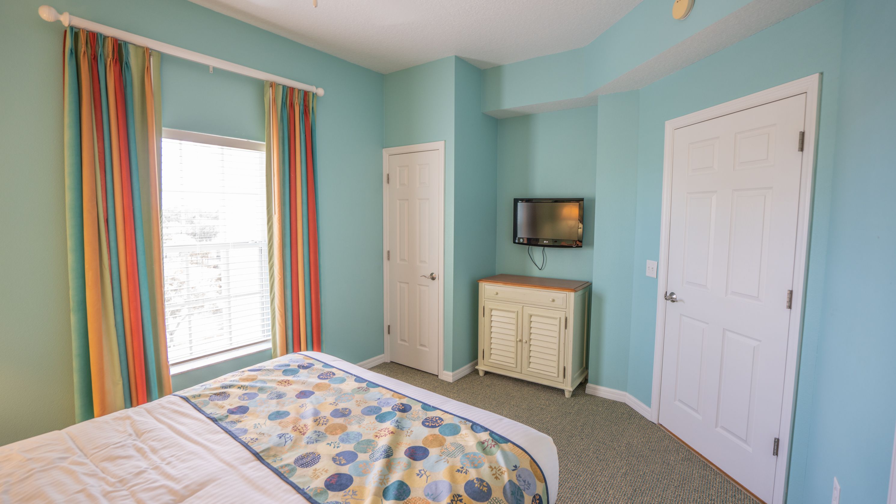Une chambre à coucher avec un lit, une commode, un téléviseur fixé au mur et deux portes.
