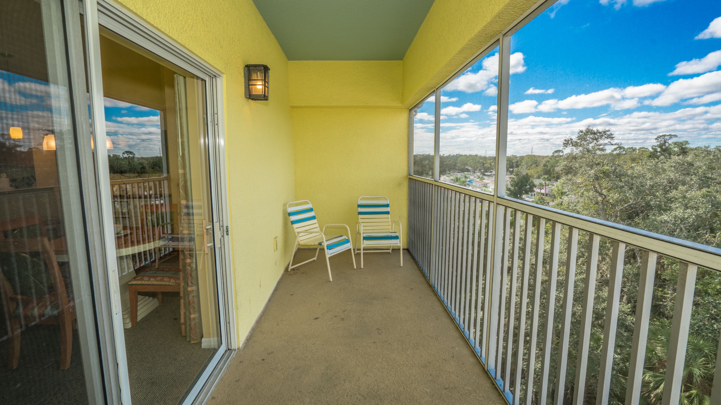 A balcony with two striped chairs, a glass door, and a view of trees and houses.