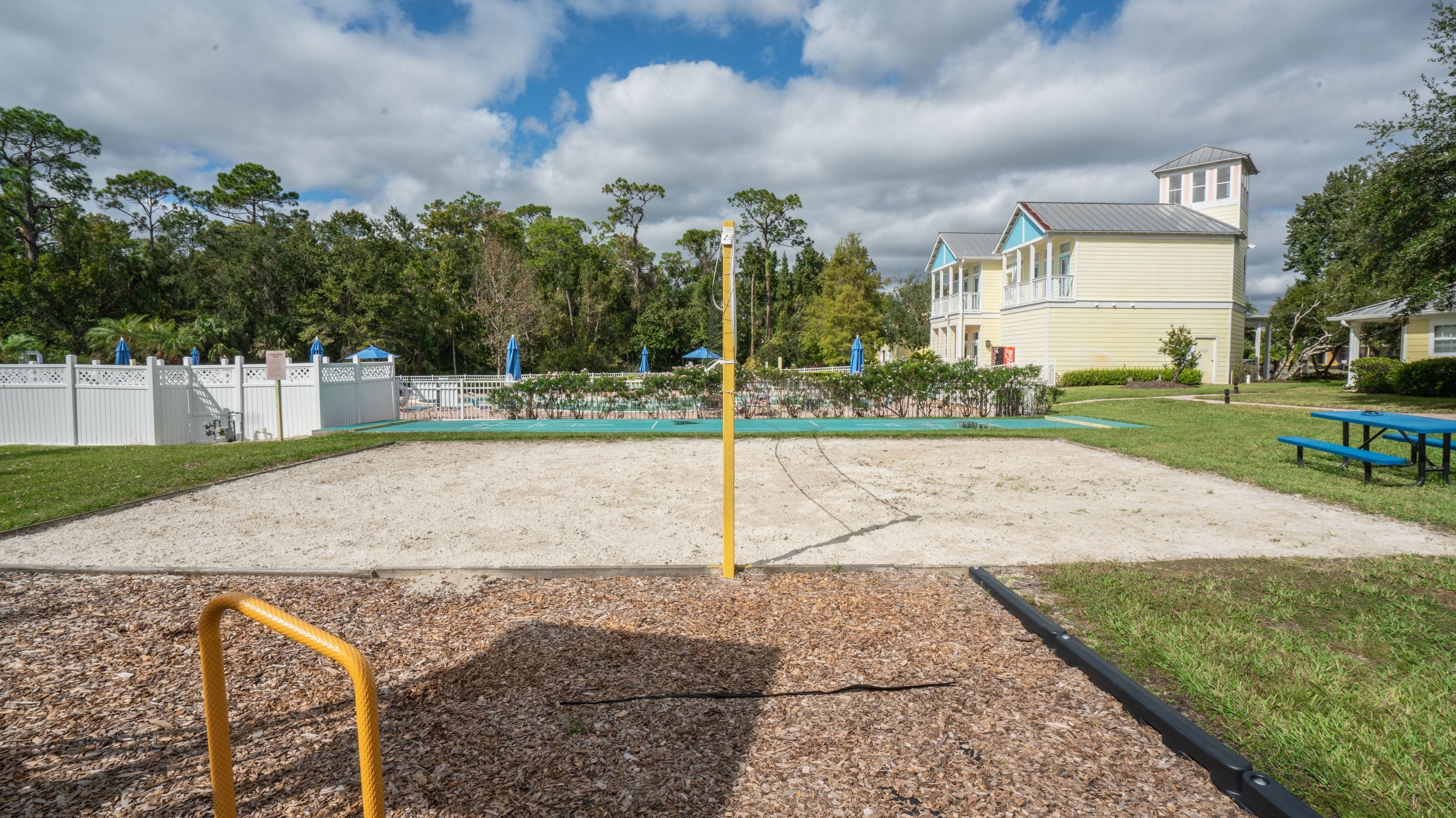 A sandy volleyball court with a net, surrounded by a white fence and trees.