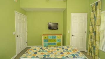 Quarto com paredes verdes, cama com colcha colorida, cómoda com várias gavetas, televisão na parede e janela com cortinas estampadas.