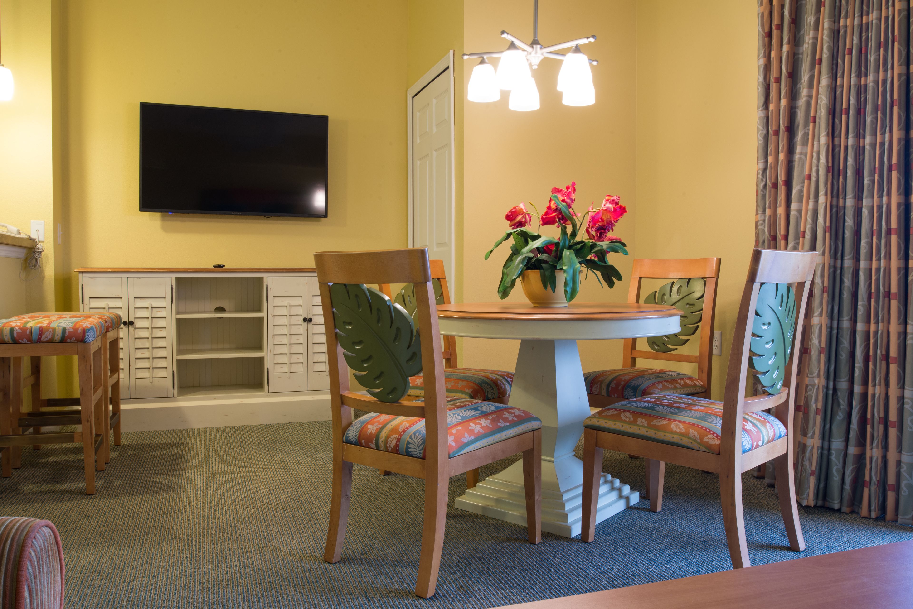 A dining area with a round table, chairs with leaf-patterned backs, a flat-screen TV mounted on the wall, and a vase with flowers.