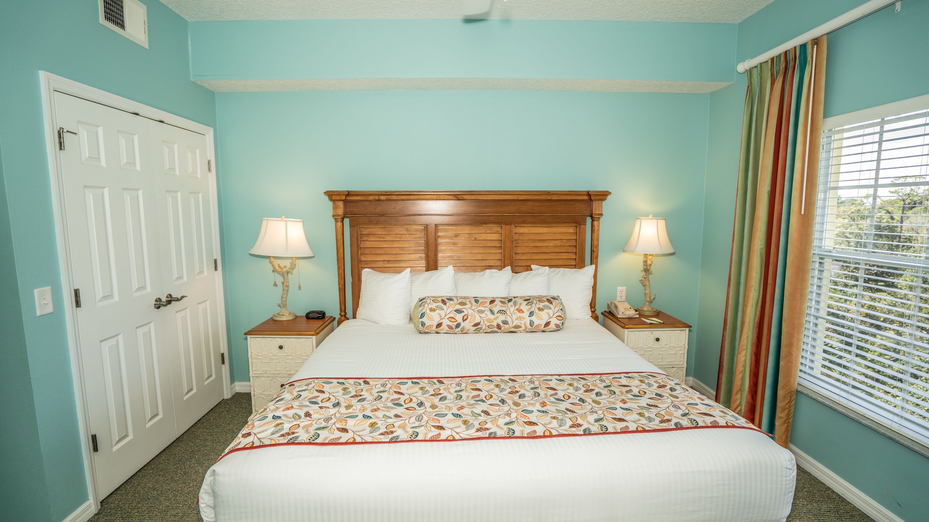 A bedroom with a large bed, two bedside tables with lamps, a wooden headboard, and a window with blinds.