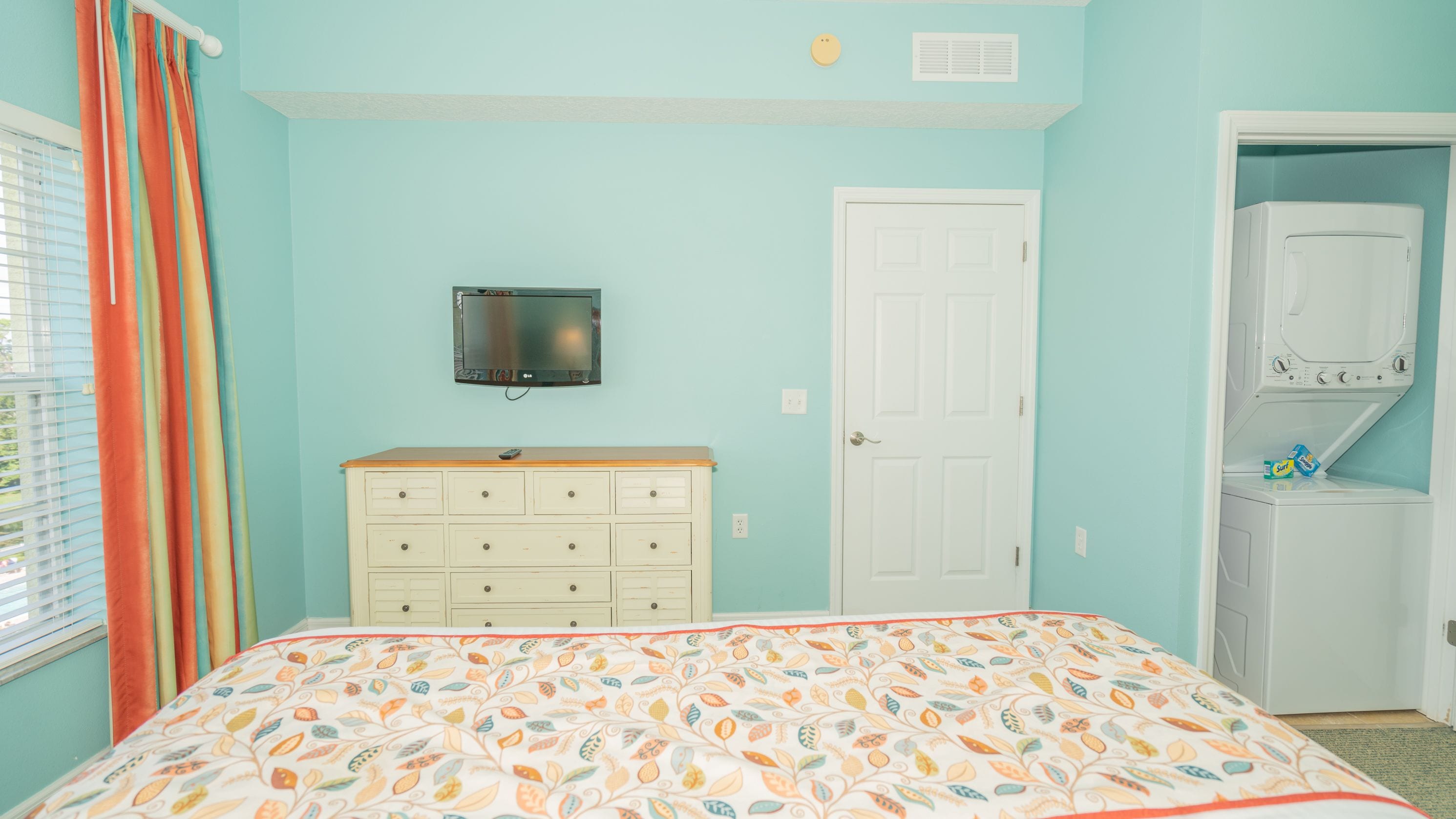 A bedroom with a bed, dresser, television, closet, and window with curtains.