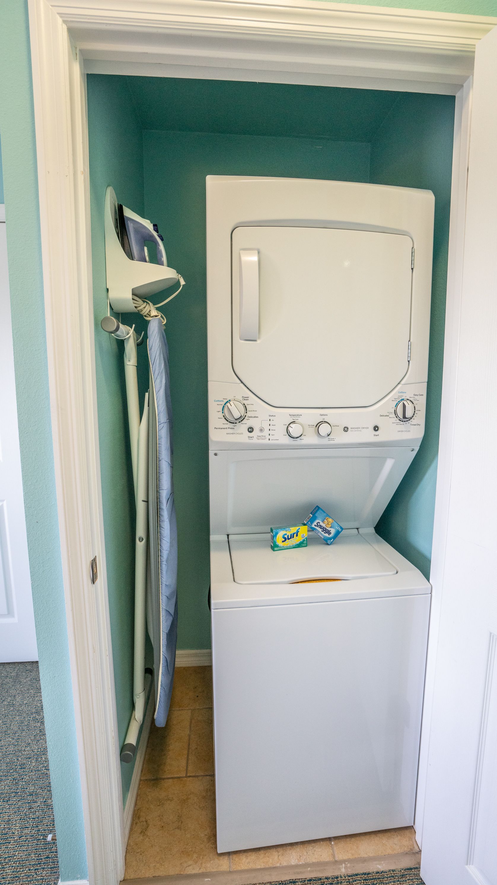 Une petite buanderie équipée d’un lave-linge et d’un sèche-linge à chargement frontal, d’un étendoir et d’une petite étagère où sont rangées les boîtes de lessive.