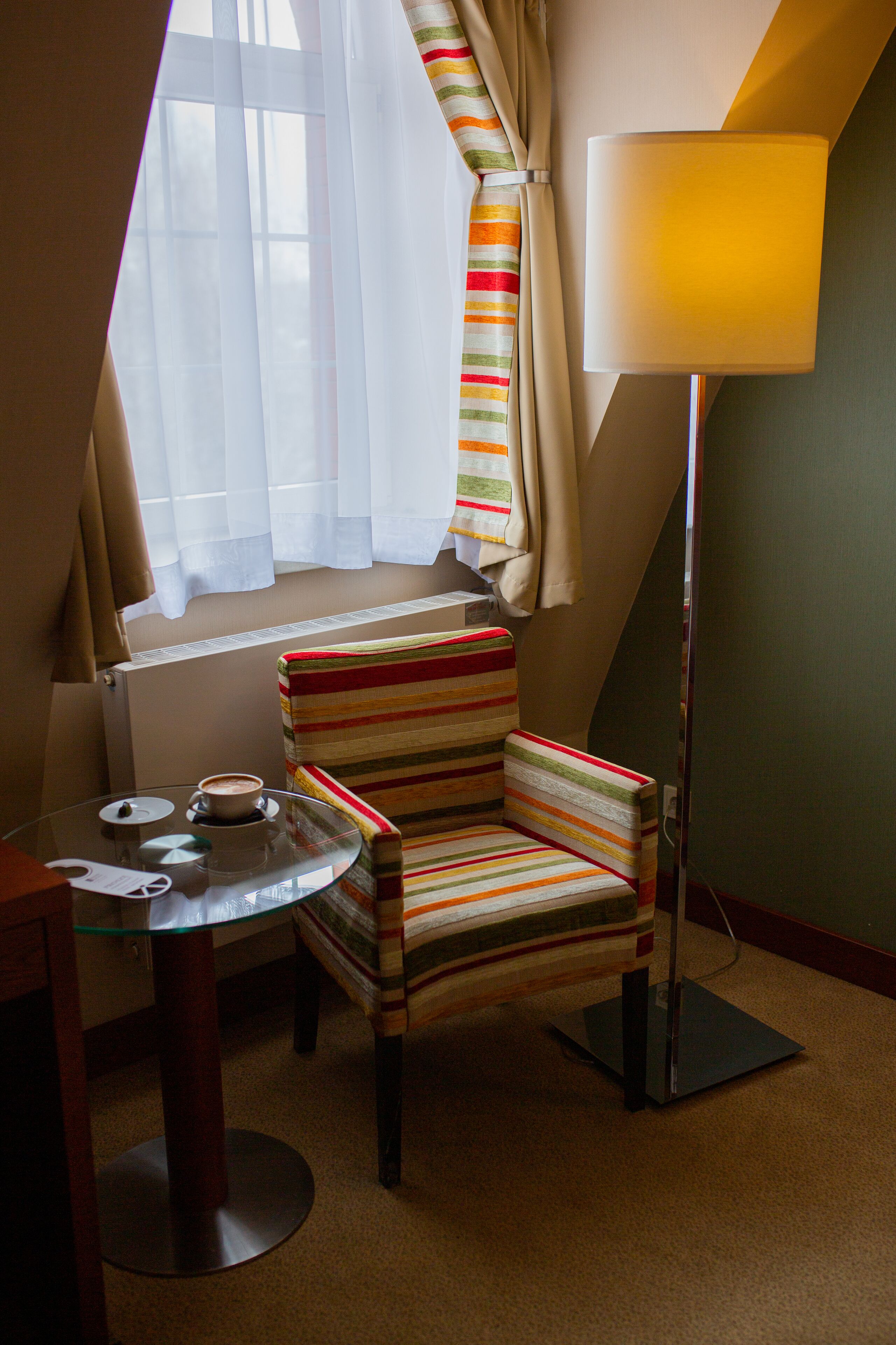 A cozy room with a striped armchair, a glass table with a cup and saucer, and a standing lamp.
