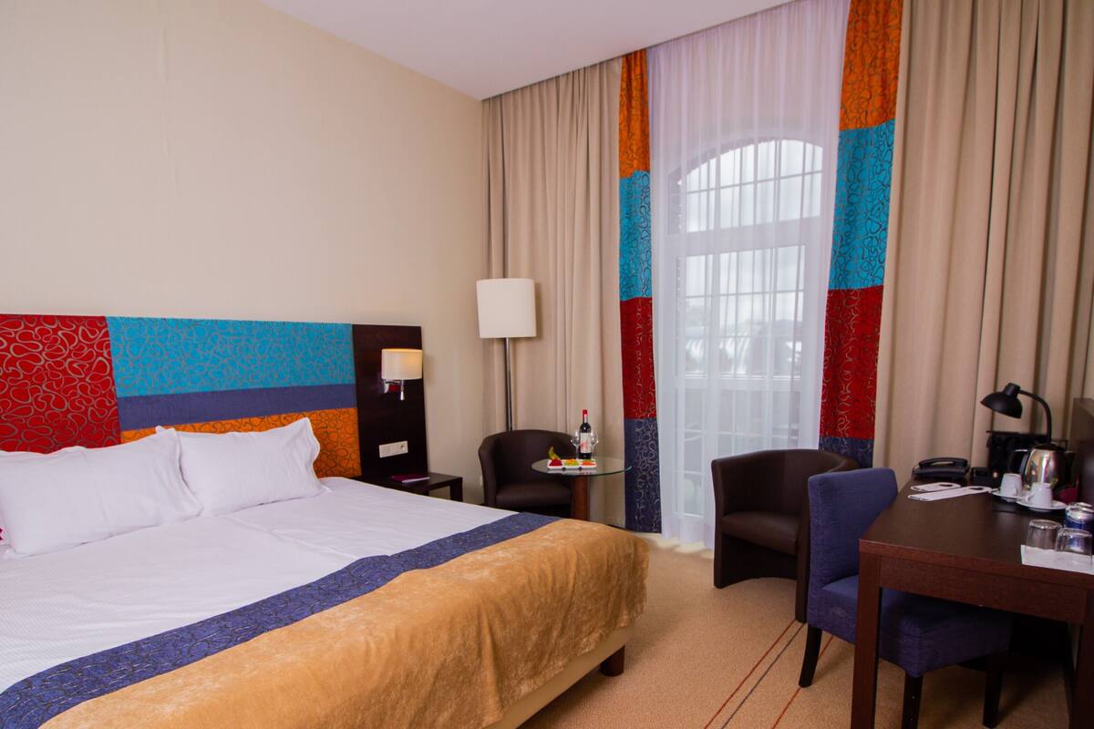 Una habitación de hotel con una cama grande, un escritorio, una silla y un ventanal con cortinas.