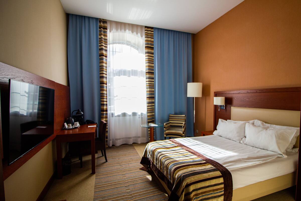 Una habitación de hotel con cama, escritorio, silla, televisor y ventana con cortinas.