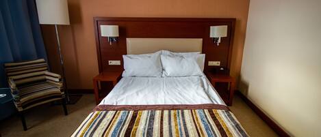 Habitación de hotel con una cama grande, dos lámparas de noche, una manta a rayas, una silla a rayas y una lámpara de pie.