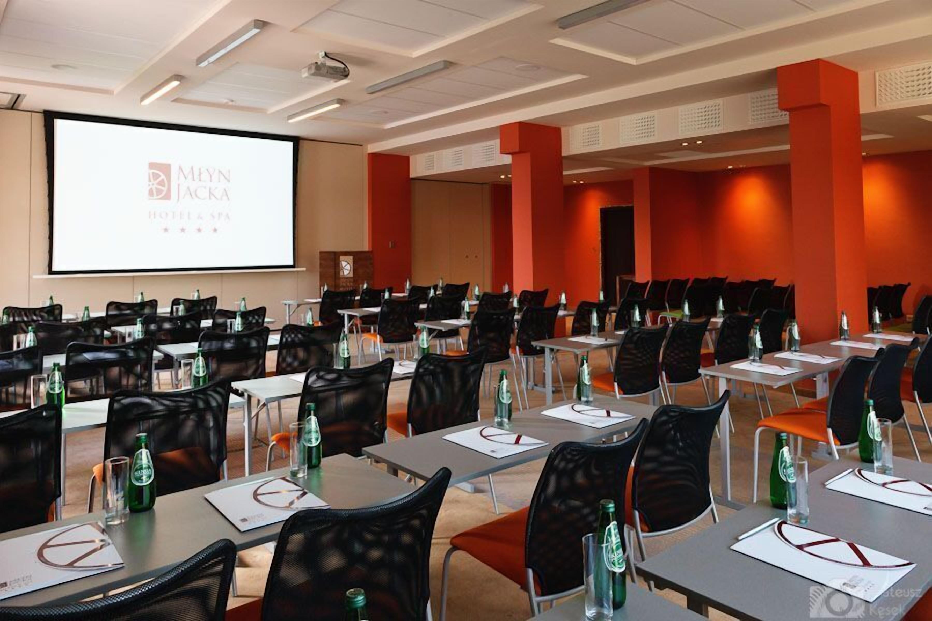 A conference room with multiple tables and chairs, a projector screen displaying the hotel name, and a stage area.