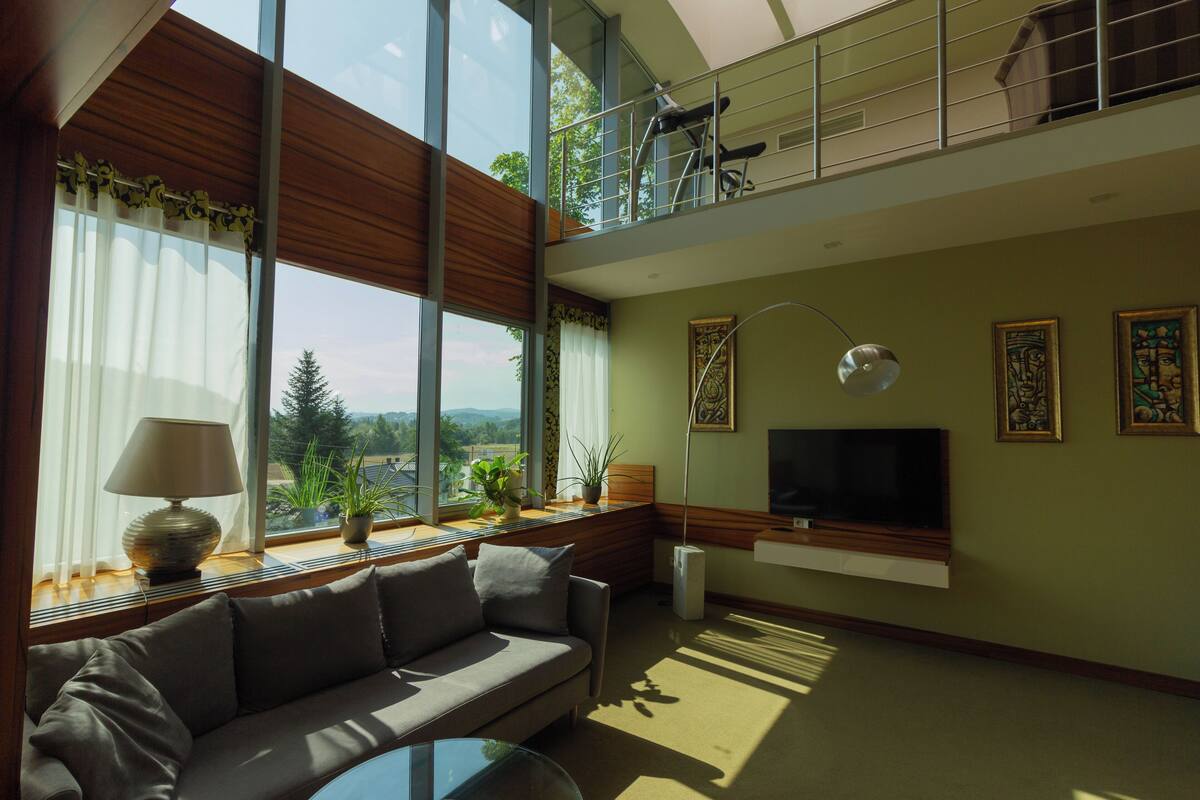 Una sala de estar moderna con un sofá, un televisor montado en la pared y grandes ventanas con vistas al exterior.